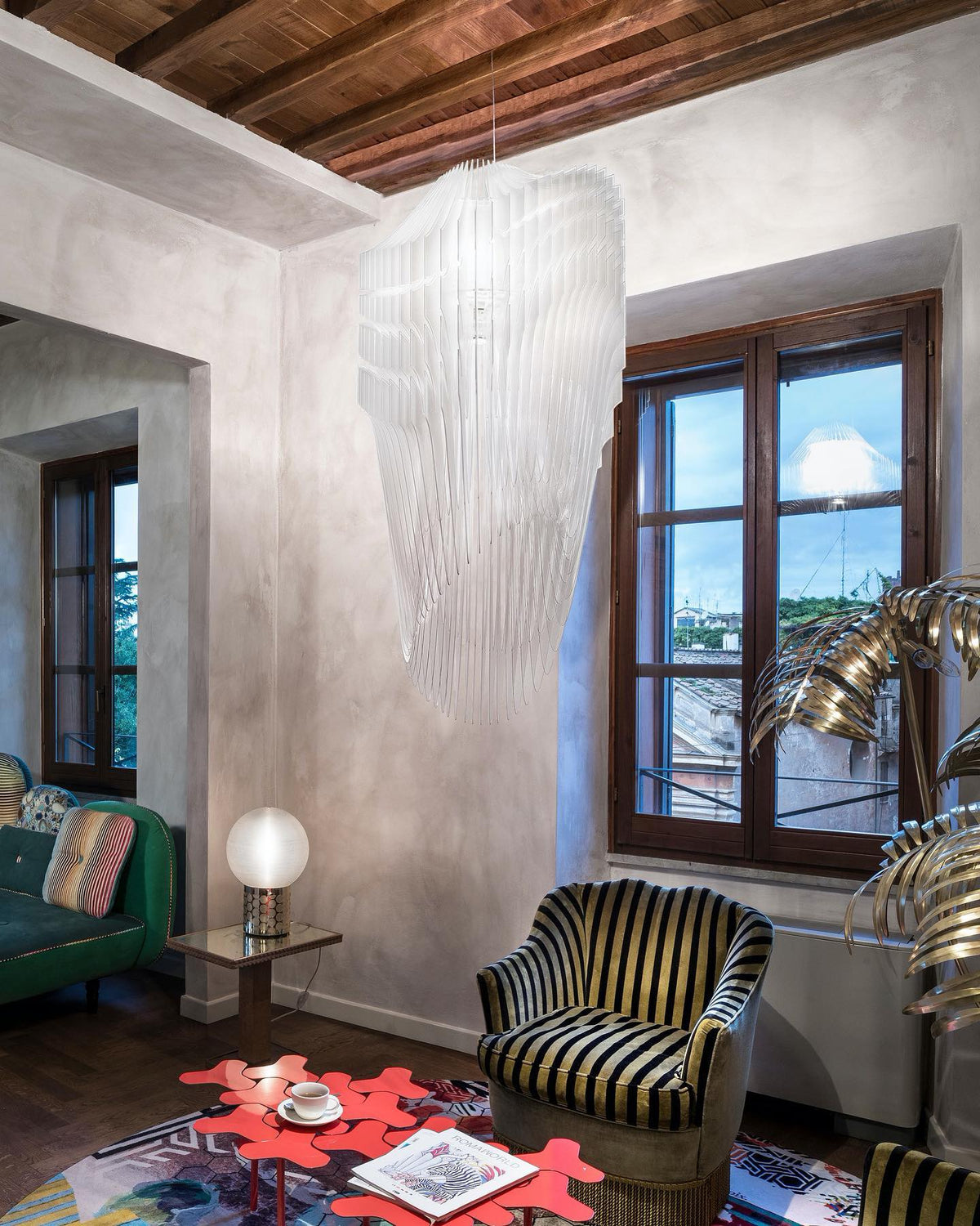 A clear acrylic fringe pendant from Clérachic hangs in a corner of a room with a beamed ceiling, a striped armchair and a red coffee table.