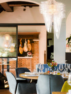 Tall view of a clear fringe pendant from Clérachic above a restaurant table set with wine glasses, with hanging hams beyond.