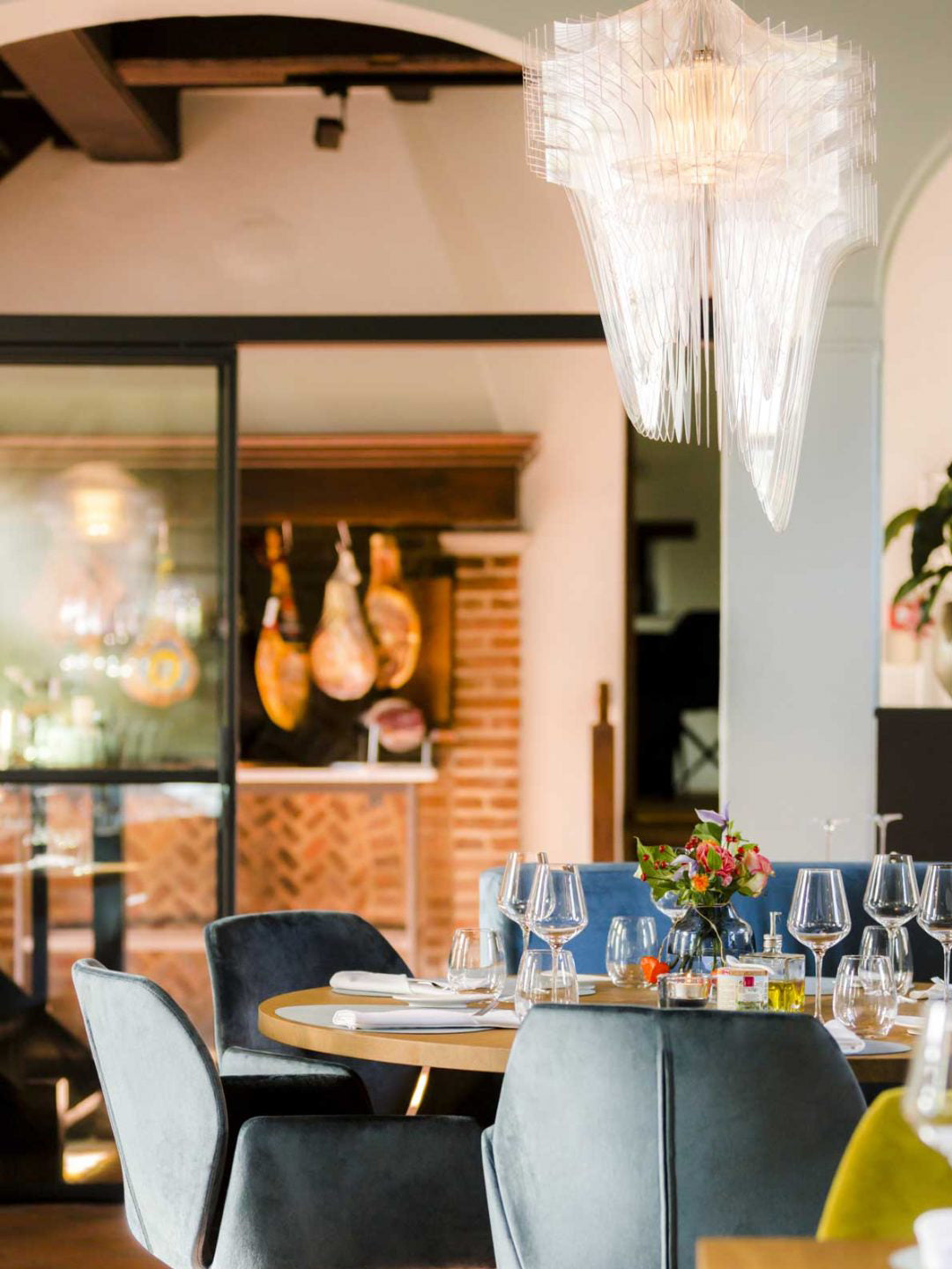 Tall view of a clear fringe pendant from Clérachic above a restaurant table set with wine glasses, with hanging hams beyond.