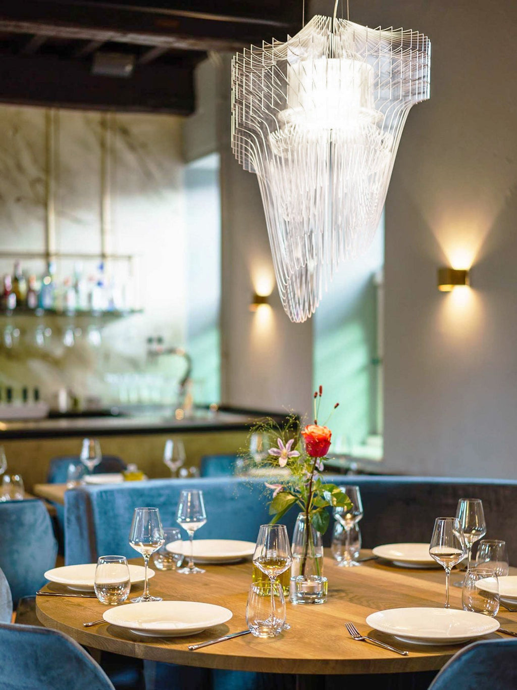 A clear acrylic fringe pendant from Clérachic hangs above a round dining table with glasses and flowers, with brass wall lights behind.