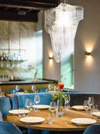 A clear acrylic fringe pendant from Clérachic hangs above a round dining table with glasses and flowers, with brass wall lights behind.