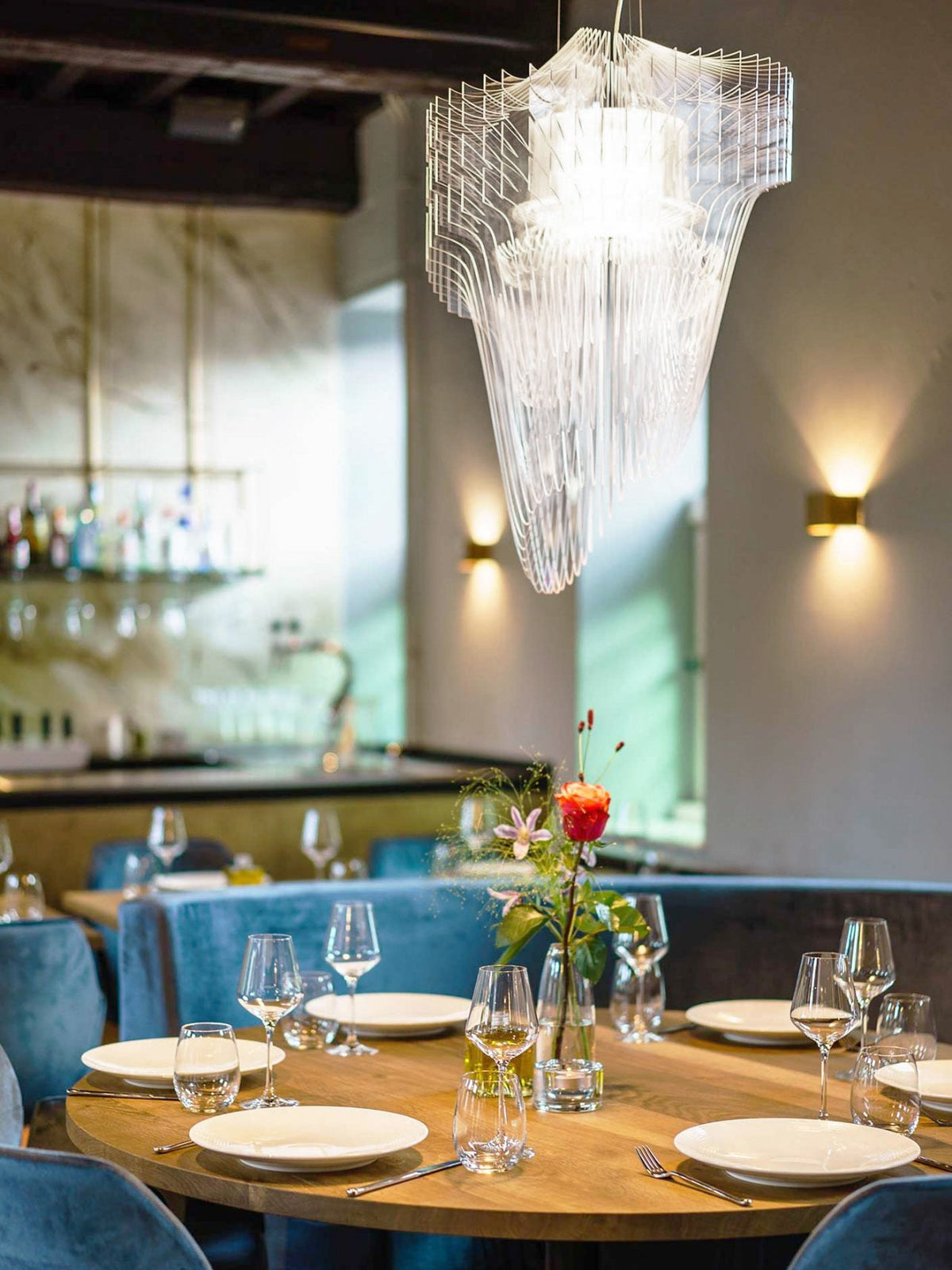 A clear acrylic fringe pendant from Clérachic hangs above a round dining table with glasses and flowers, with brass wall lights behind.