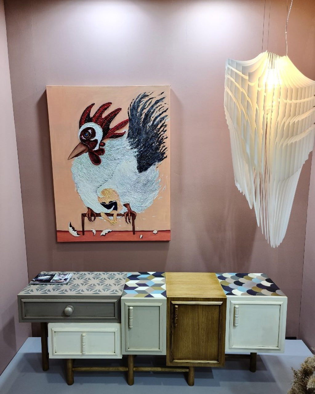 An ivory fringe light hangs on a mauve wall beside a painting of a hen, above a patterned sideboard, in a Clérachic setting.