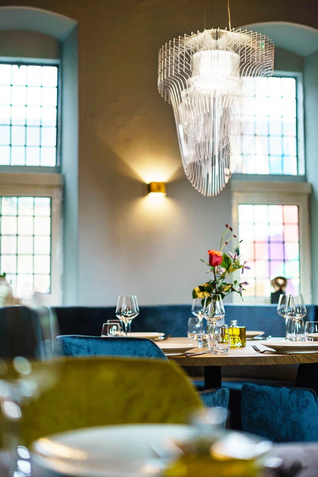 A clear acrylic fringe pendant from Clérachic hangs beside windows and a brass wall light, above a restaurant table with flowers.
