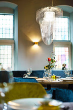 A clear acrylic fringe pendant from Clérachic hangs beside windows and a brass wall light, above a restaurant table with flowers.