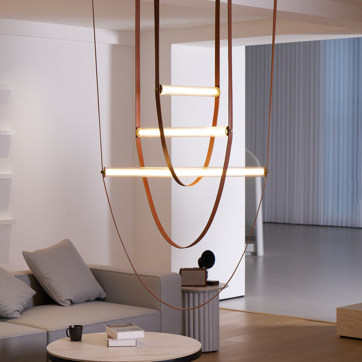 Three light bars on looping leather straps hang over a grey sofa in a bright living room, shown on this Clérachic pendant.