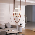 Tan leather straps curve down to three horizontal light bars of different lengths, a Clérachic pendant seen beside a grey sofa.