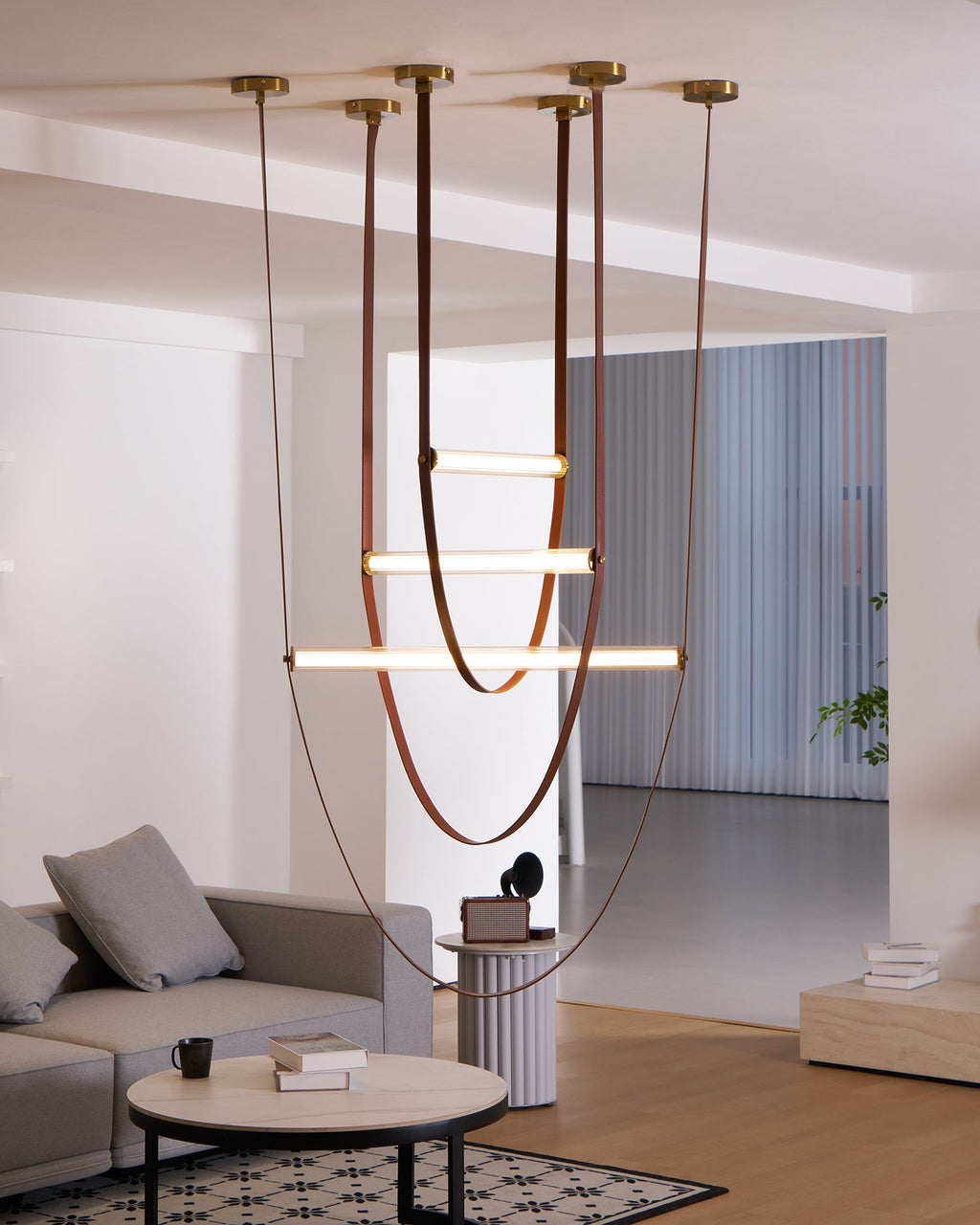 Wide living-room view of this Clérachic pendant, its leather loops falling from brass canopies past a grey sofa and a small side table.
