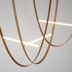 Looped tan leather straps cross two lit bars, shown close up on a plain grey ceiling and wall on this Clérachic pendant.
