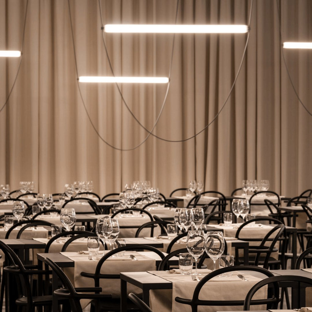 This Clérachic pendant's long light bars hang in front of a pleated curtain above a restaurant dining room with set tables.
