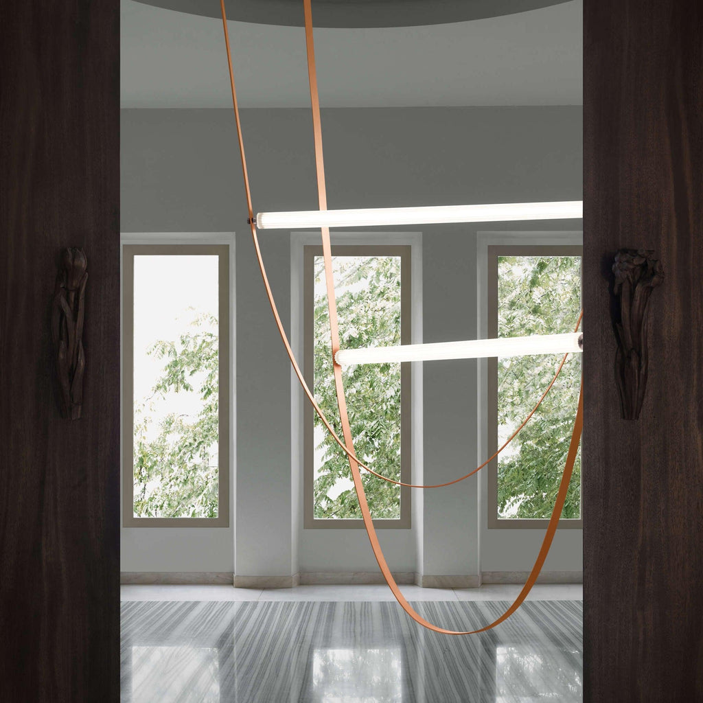 Tan leather strap loops and two horizontal light bars from this Clérachic pendant hang between dark wooden panels and tall windows.