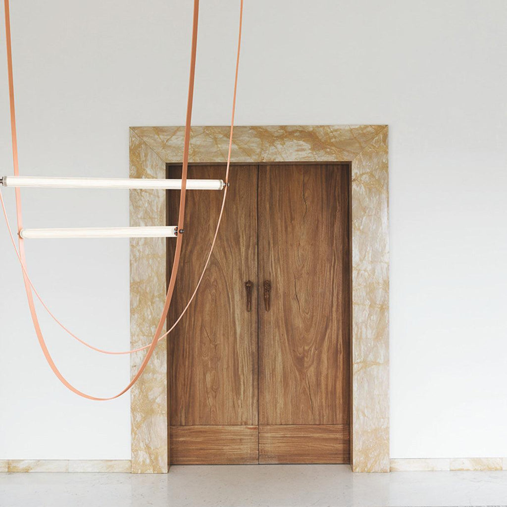 Leather straps and two horizontal bars from this Clérachic pendant hang in front of a white wall and a double timber door.
