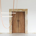 Leather straps and two horizontal bars from this Clérachic pendant hang in front of a white wall and a double timber door.