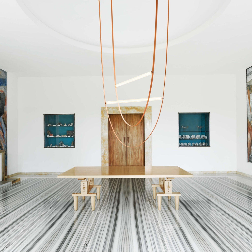 A Clérachic pendant with three leather loops hangs in a round white room with a wooden door and a long stone-topped table.