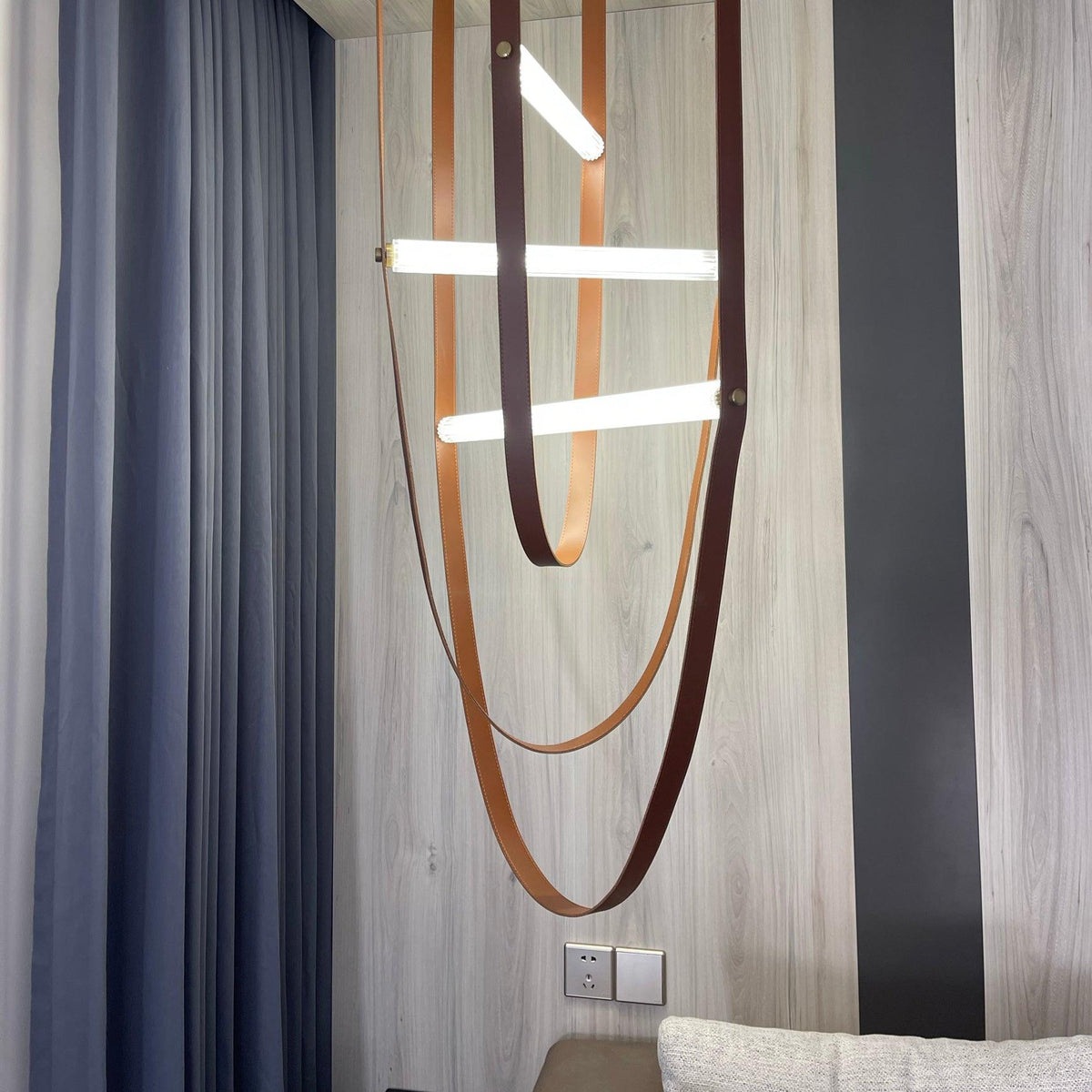 Leather loops and angled light bars from this Clérachic pendant hang against a grey wall and striped blue curtain.