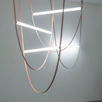 Three light bars on copper-toned leather loops hang beside a black version, a Clérachic pendant set against a pale grey wall.