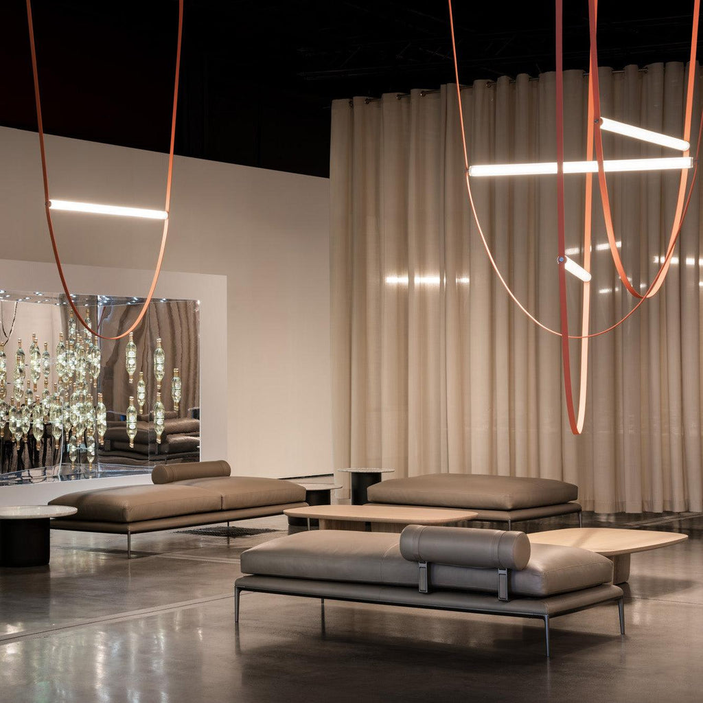 Leather loops and bars from this Clérachic pendant hang in front of cream curtains in a lounge with low grey sofas.
