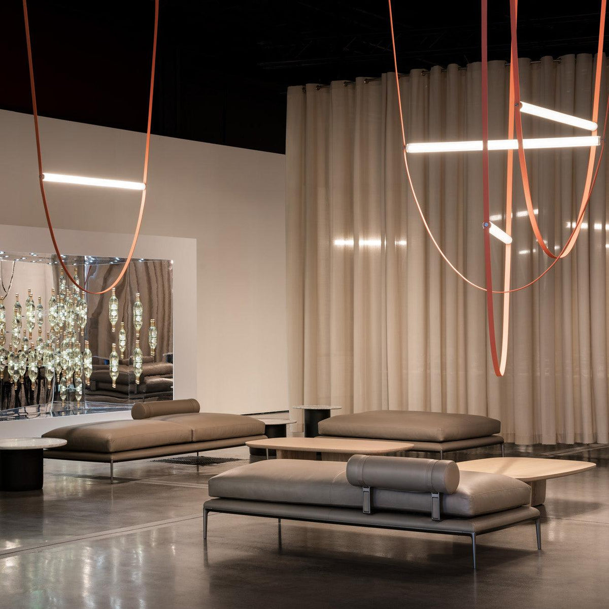 Leather loops and bars from this Clérachic pendant hang in front of cream curtains in a lounge with low grey sofas.