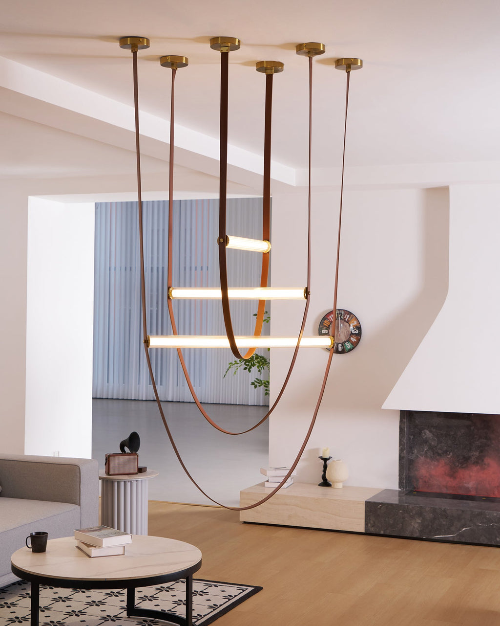 Six brass canopies feed this Clérachic pendant's tan leather loops and three bars, in a bright room with a fireplace and wall clock.