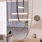 Three lit bars on tan leather loops from this Clérachic pendant, with a round wall clock and a grey sofa behind.