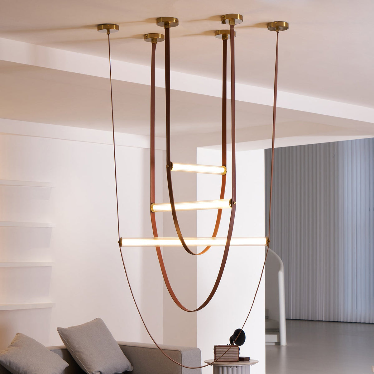 This Clérachic pendant seen from a low angle, with three lit bars hung from six canopies in a white room with a sofa and grey blinds.