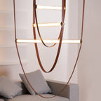 Looped tan leather straps and three lit bars from this Clérachic pendant, seen close up with shelves glowing behind.