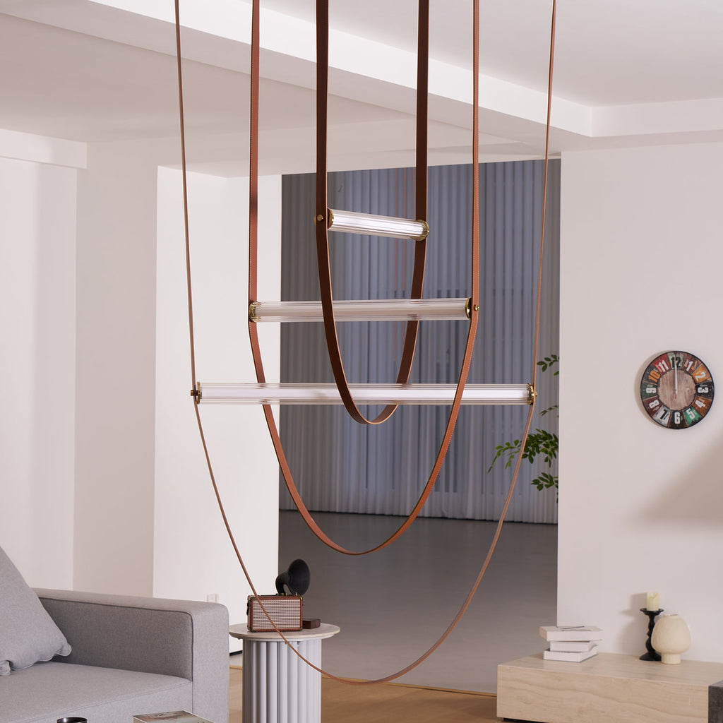 A Clérachic pendant with three loops and bars of different lengths hangs in a white room with a wall clock and grey sofa.