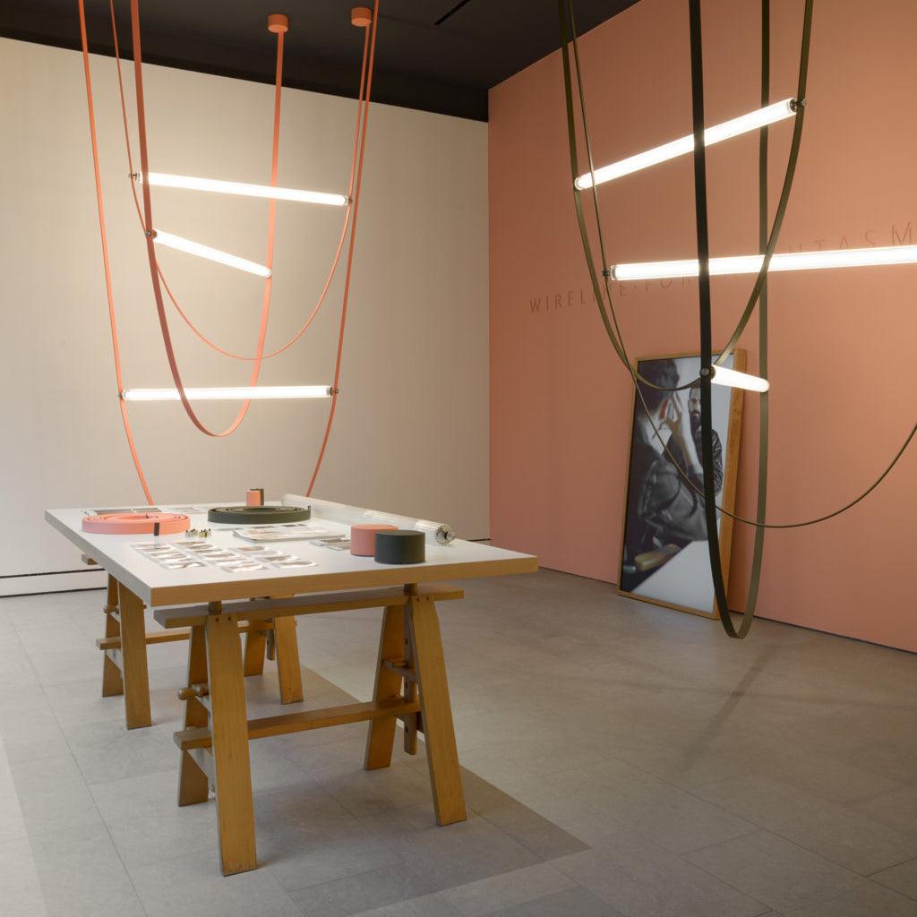 Tan and dark leather loops with glowing bars from this Clérachic pendant, in a gallery space with a pink wall and wooden trestle table.