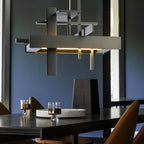 Close view of a dark bronze-toned geometric Clérachic pendant with layered bars and a warm glow, above a dark table and a blue wall.