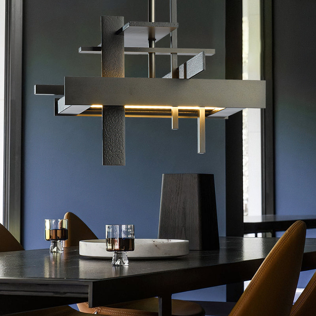 Close view of a dark bronze-toned geometric Clérachic pendant with layered bars and a warm glow, above a dark table and a blue wall.