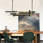 A wide dark Clérachic pendant with layered, offset bars glows warm above a timber dining table with dark green chairs and a large painting.