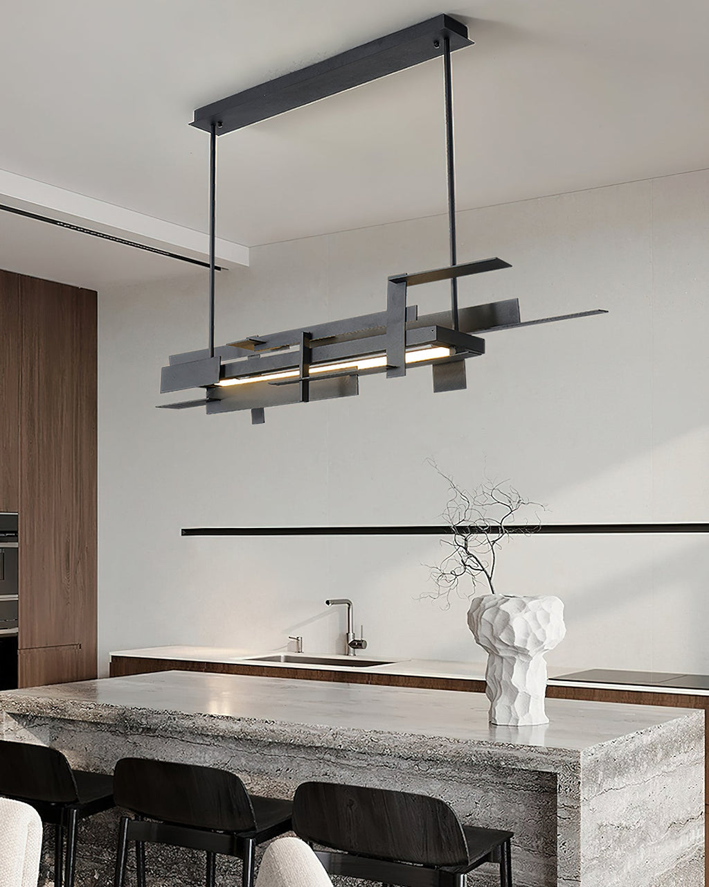 A slim black linear Clérachic pendant hangs above a stone-topped kitchen island with black chairs and a sculptural white vase.