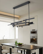 A linear black Clérachic pendant hangs above a black dining table in a white kitchen with built-in ovens and a warm ceiling glow.