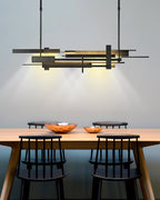 A wide dark Clérachic pendant with offset bars casts a warm glow on a timber table with copper bowls and spindle-back chairs.
