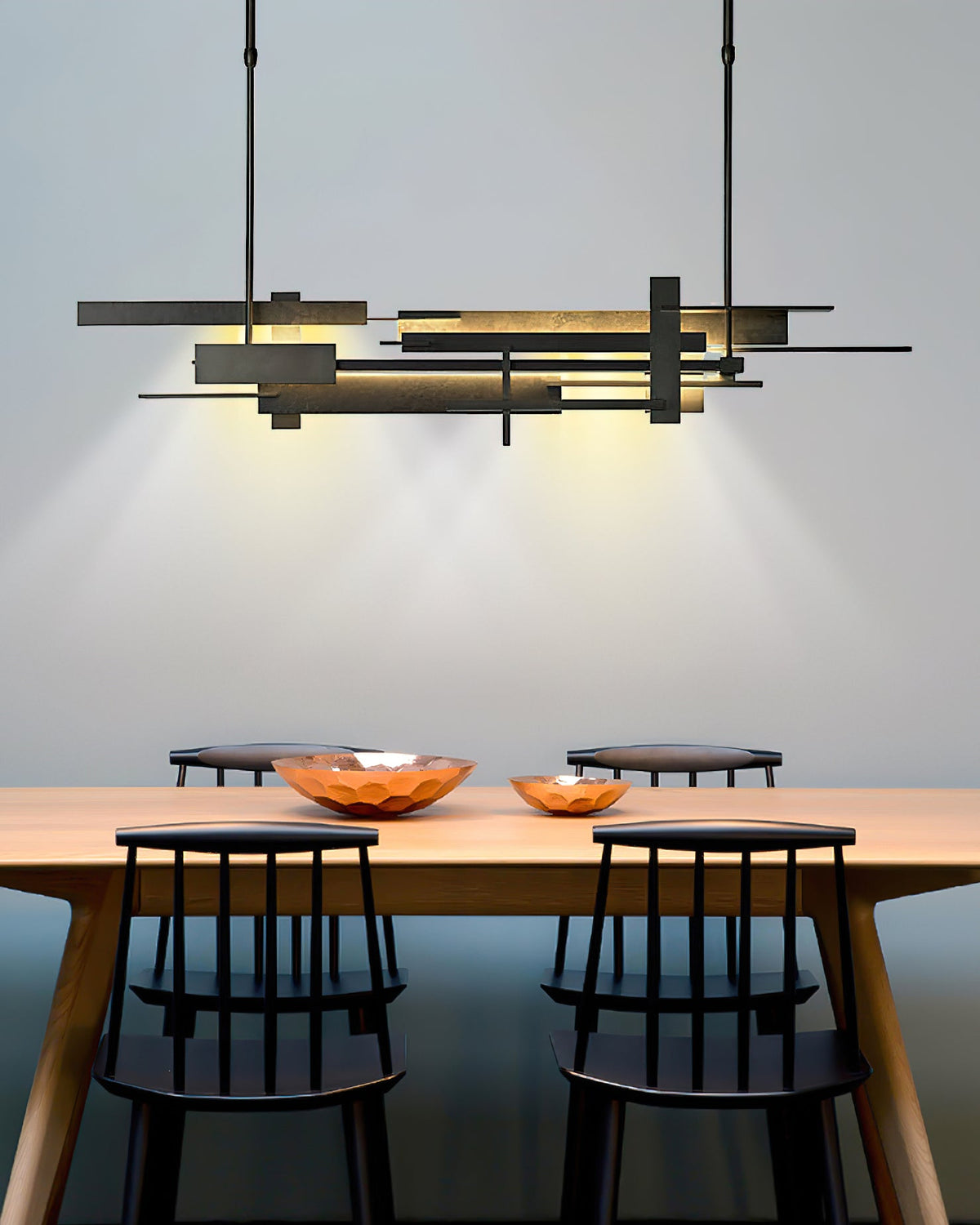 A wide dark Clérachic pendant with offset bars casts a warm glow on a timber table with copper bowls and spindle-back chairs.