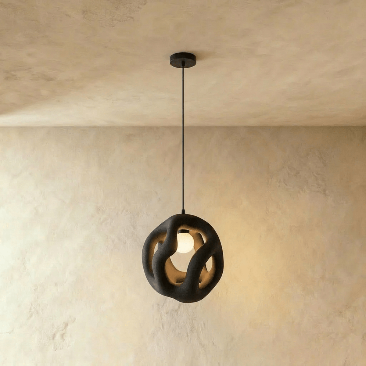 Clérachic sculptural openwork black pendant light on a slim black cord, its round bulb glowing within, against warm plaster.
