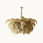 A Clérachic feather chandelier in a natural sandy tone hangs from a slim brass stem against a soft off-white backdrop.