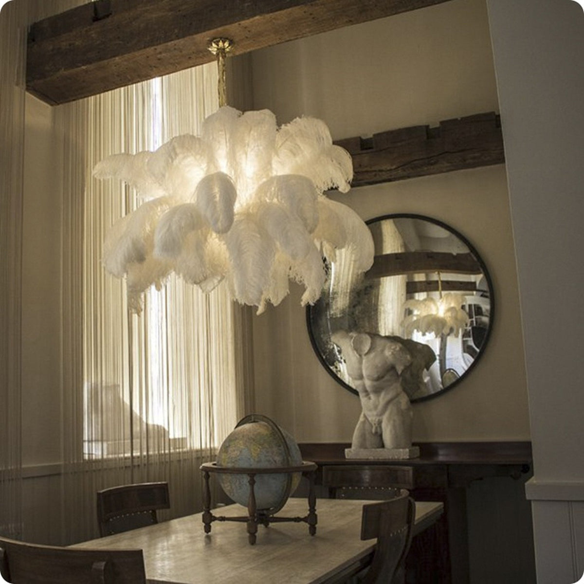 White feathers glow softly from within this Clérachic chandelier, which hangs above a wooden dining table and a round mirror.