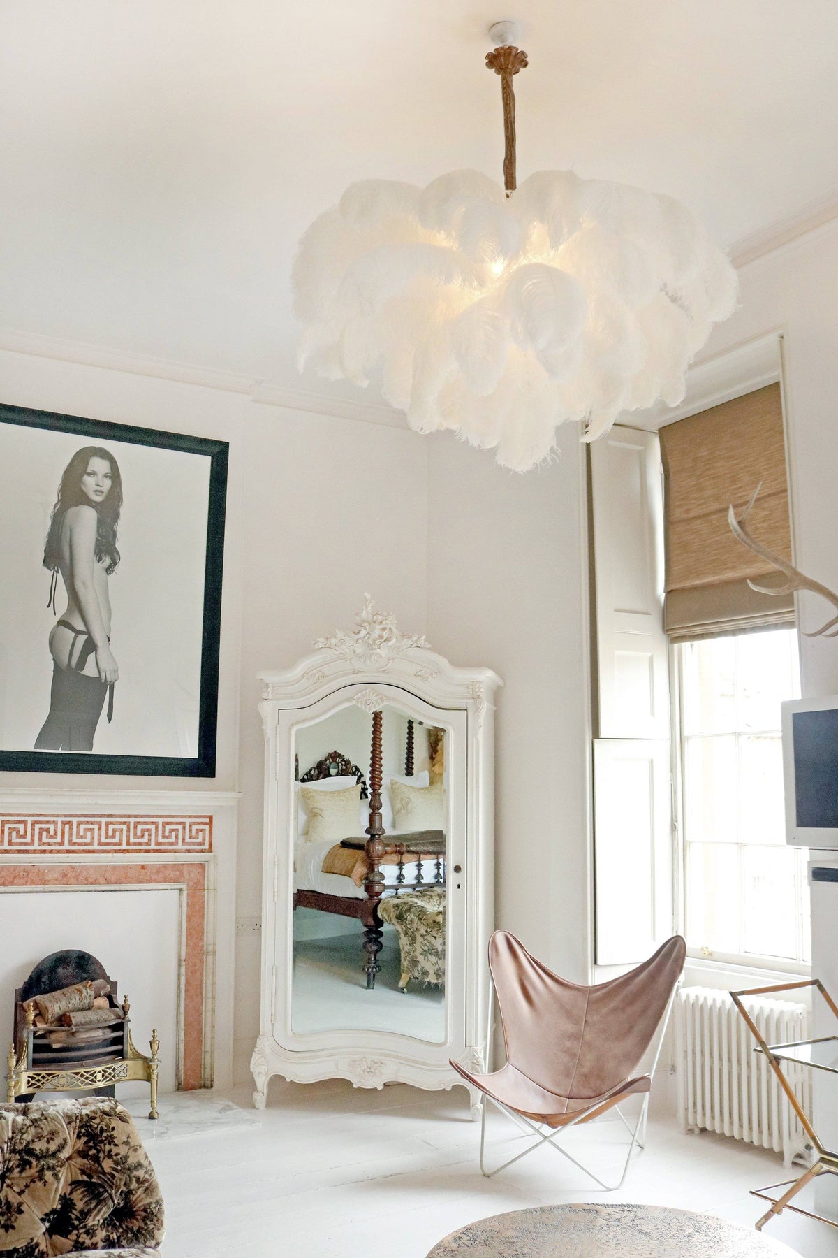 A white feather chandelier from Clérachic lit in a bright room with a pale fireplace, an armoire and a butterfly chair.