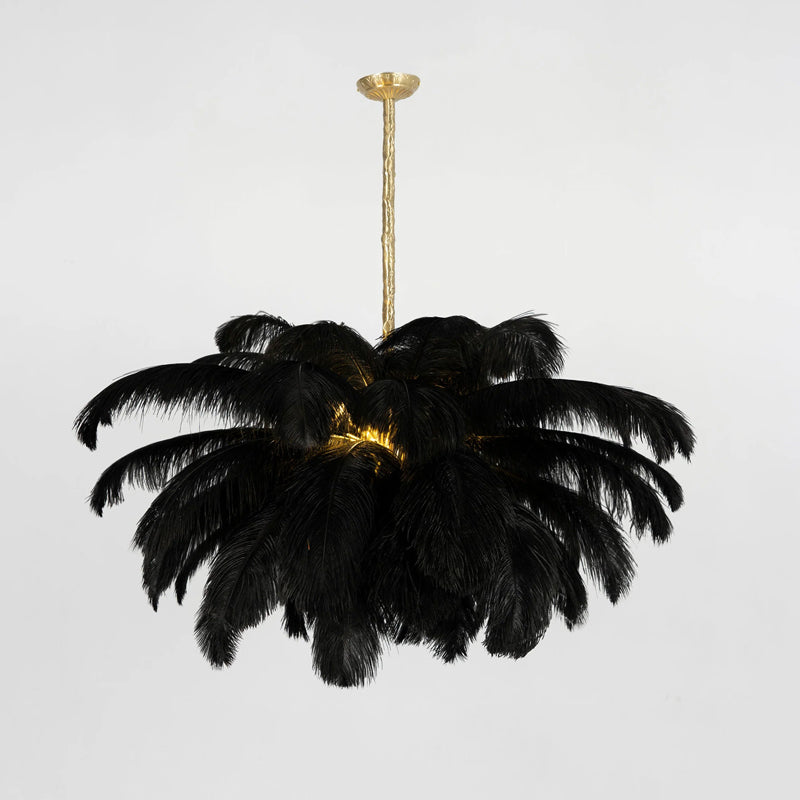 A black-feather Clérachic chandelier is lit at its core, hung from a short brass stem against a pale grey wall.