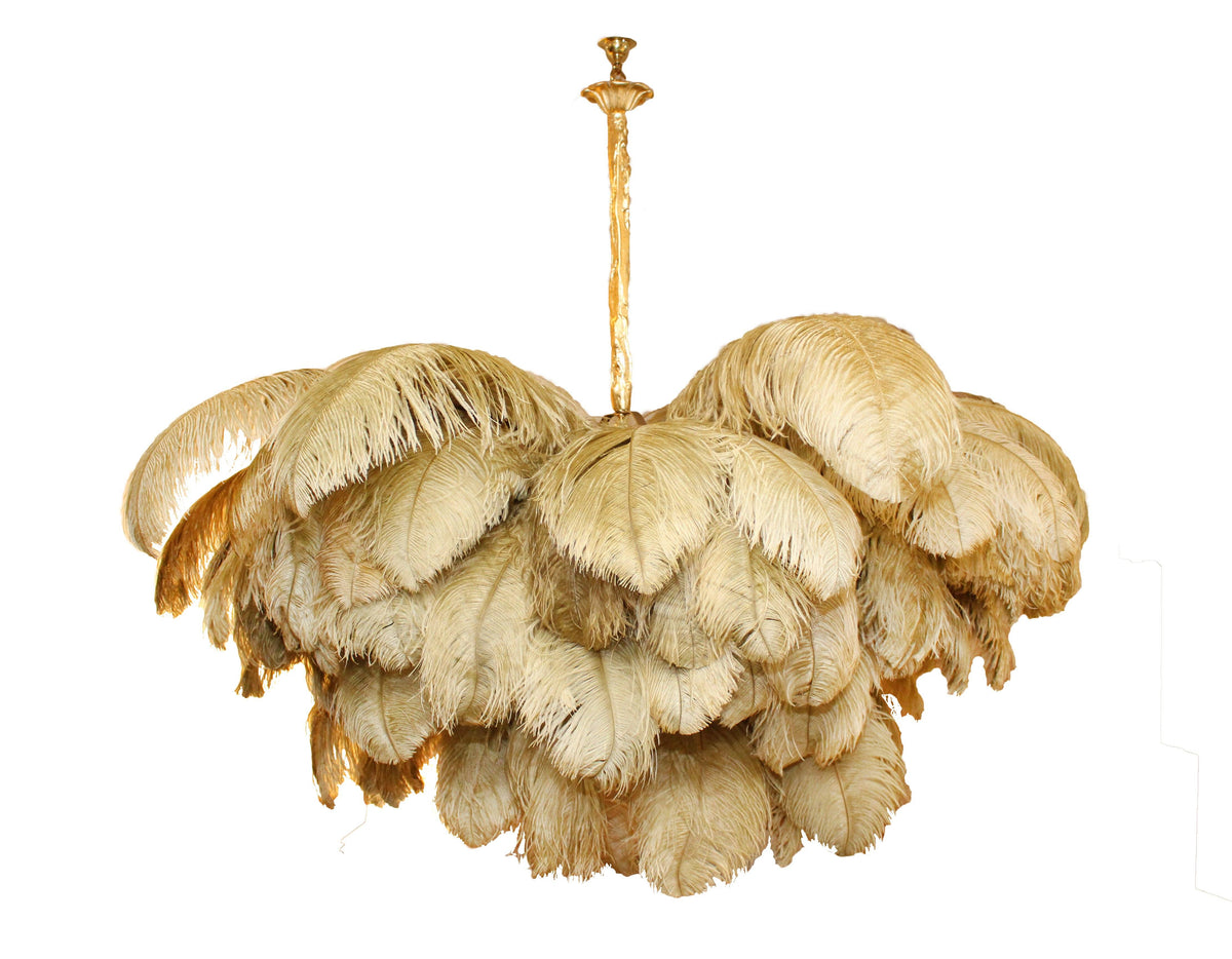 A Clérachic chandelier in gold-toned feathers hangs from a carved brass stem, shown whole on a white backdrop.