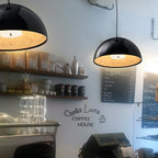 Two Clérachic glossy black dome pendant lights glowing warm over shelves of jars and coffee bags in a café.