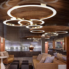 A cluster of lit gold rings, one very large, hangs below a slatted timber ceiling in a lounge with rattan seating, shown with this Clérachic pendant.