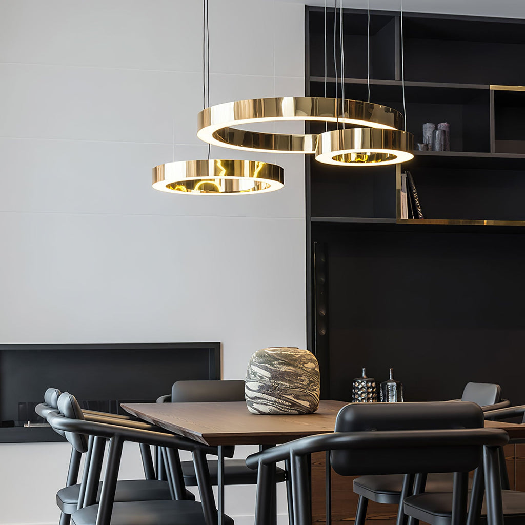 Three gold rings hang low above a walnut dining table with black chairs and a dark shelving wall, lit by this Clérachic ring pendant.