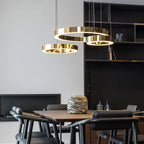 Three gold rings hang low above a walnut dining table with black chairs and a dark shelving wall, lit by this Clérachic ring pendant.