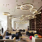 Many gold ring pendants at different heights fill the ceiling of a large open lounge with navy armchairs, in the style of this Clérachic ring pendant.