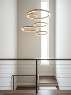 A cascade of five gold rings drops through a stairwell beside a tall narrow window, with a railing below, in a Clérachic ring pendant arrangement.