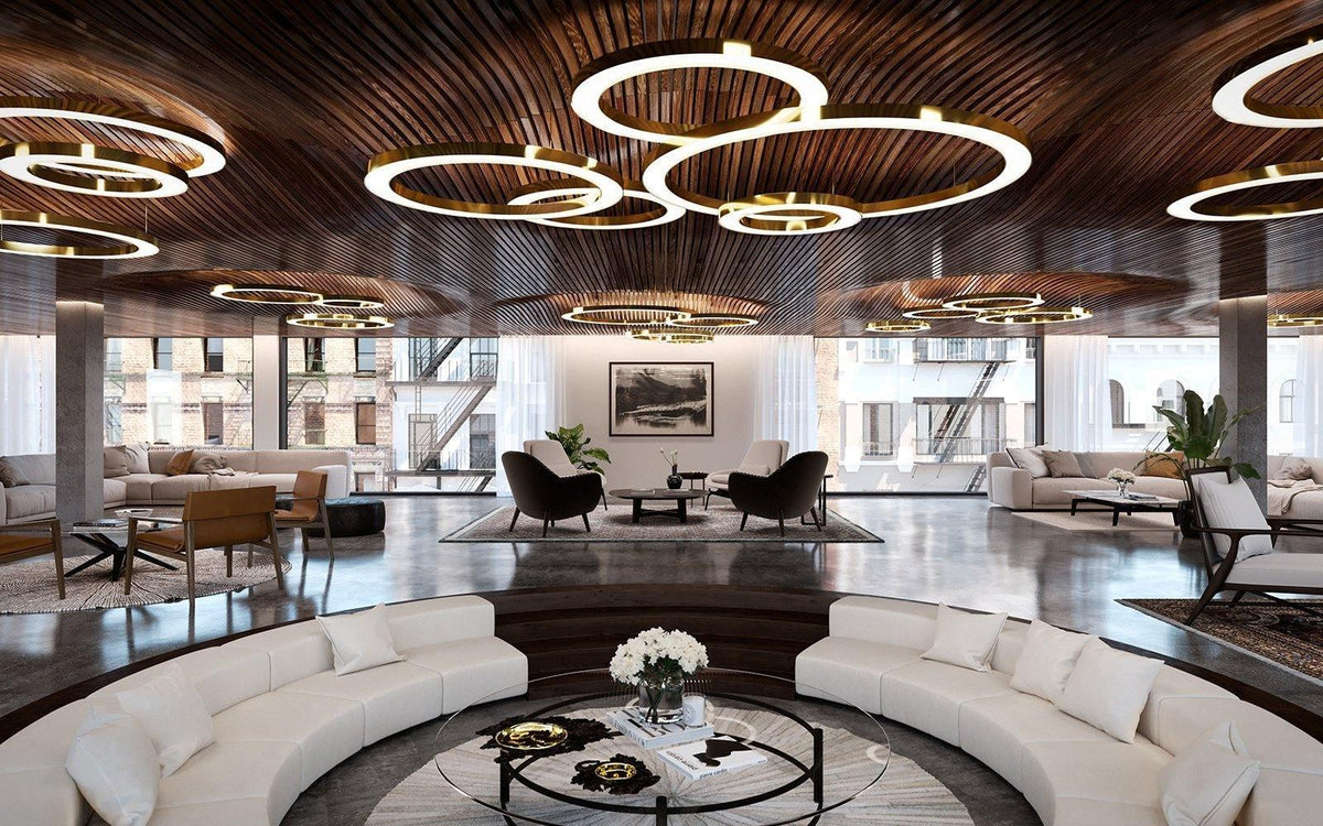 Lit gold rings hang in overlapping groups below a timber-slat ceiling in a wide lounge with curved white seating, in a Clérachic ring pendant setting.