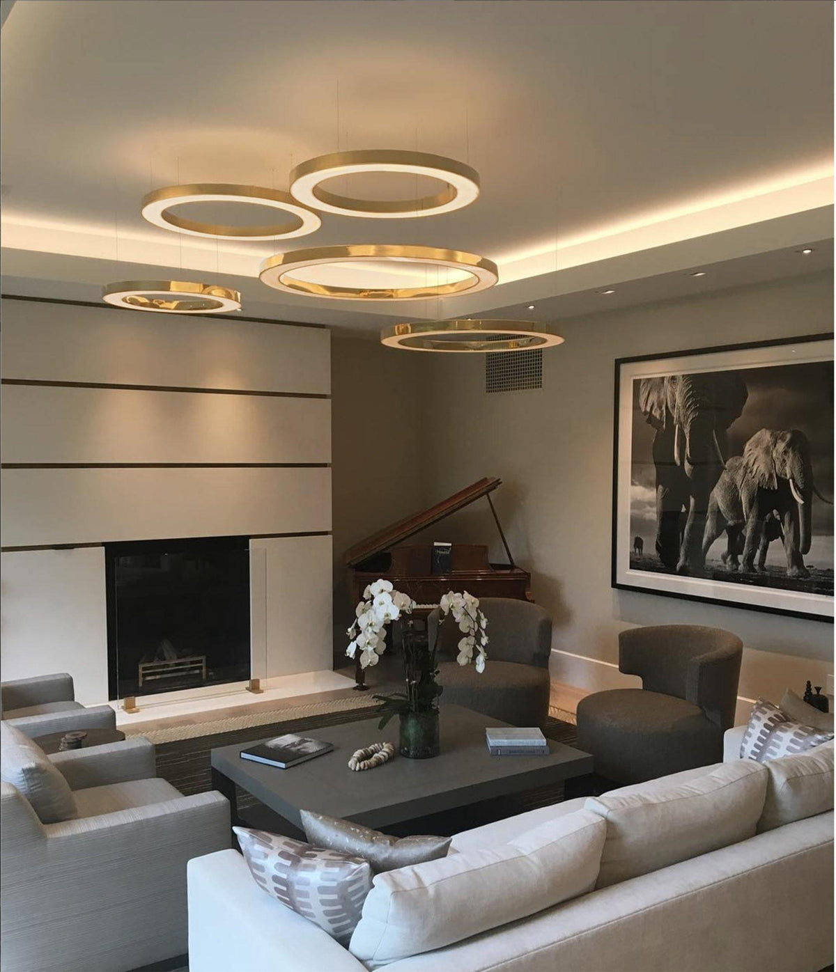 Five gold rings glow below a ceiling with a lit cove edge in a living room with a grand piano and a large framed photograph, from Clérachic.