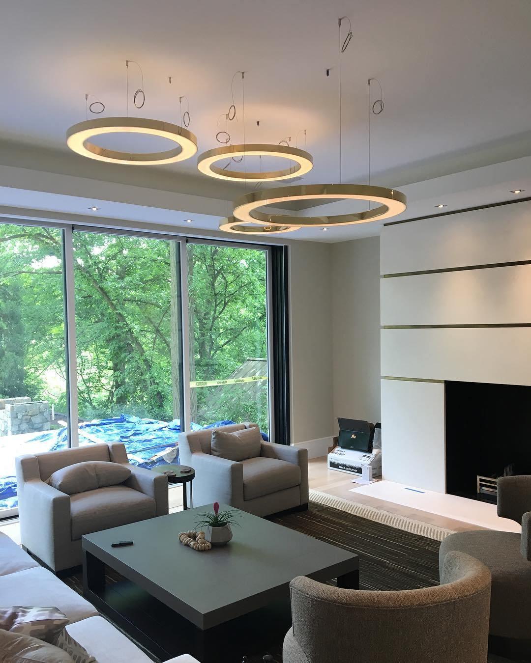 Four gold rings hang beneath tall windows onto trees in a living room with grey armchairs and a low square table, lit by this Clérachic pendant.
